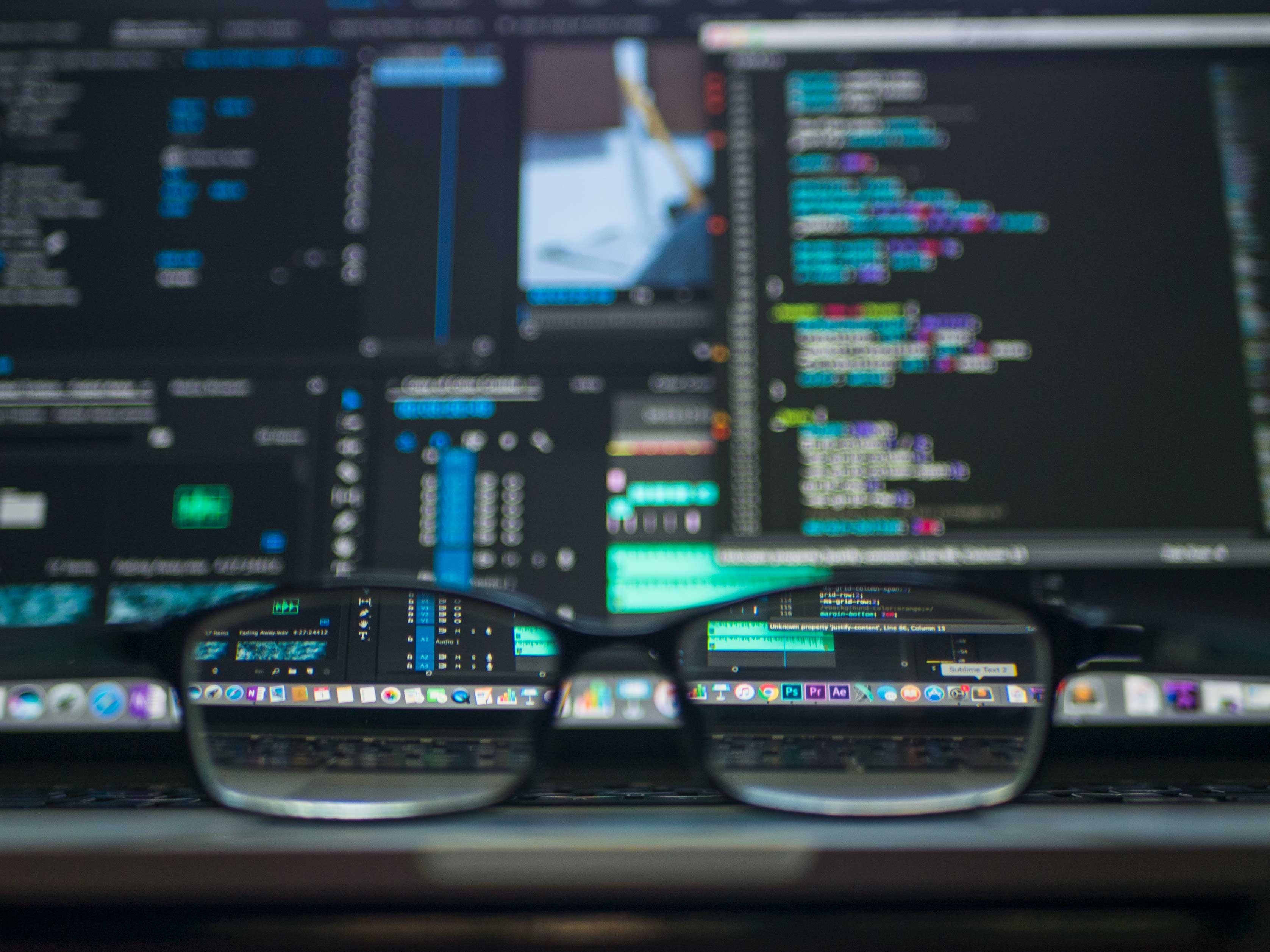 A pair of glasses sitting on a table facing two blurred screens filled with code